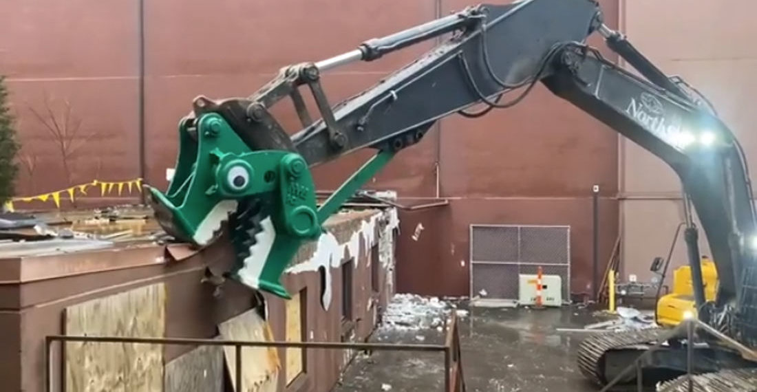 An Excavator Painted Like A Dinosaur Head With Googly Eyes Eats A Building