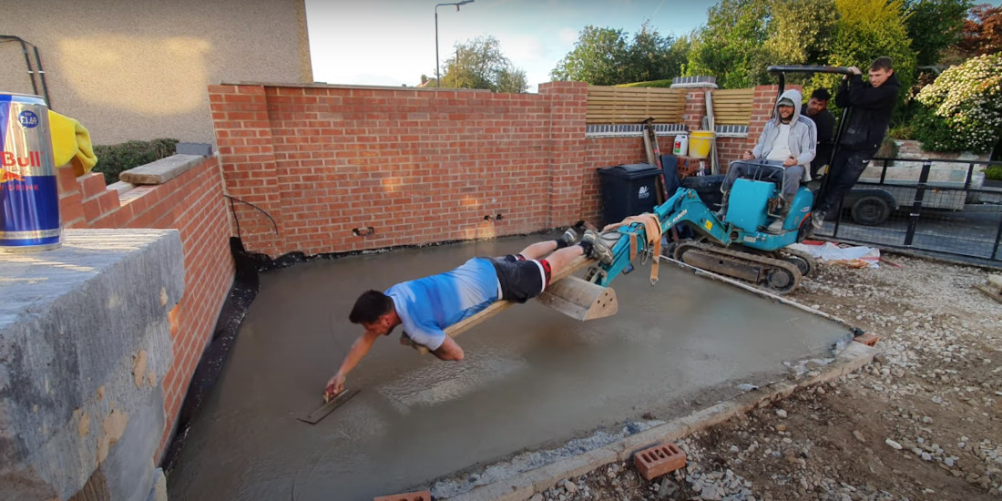 Man Laying On Extended Digger Arm Smooths Fresh Concrete