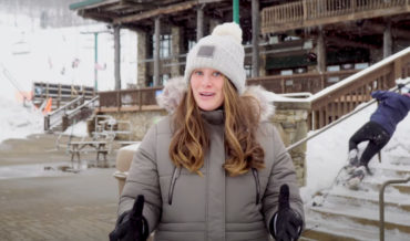Woman Delivers Ski Resort Snow Report While Skier Struggles Down Stairs In Background