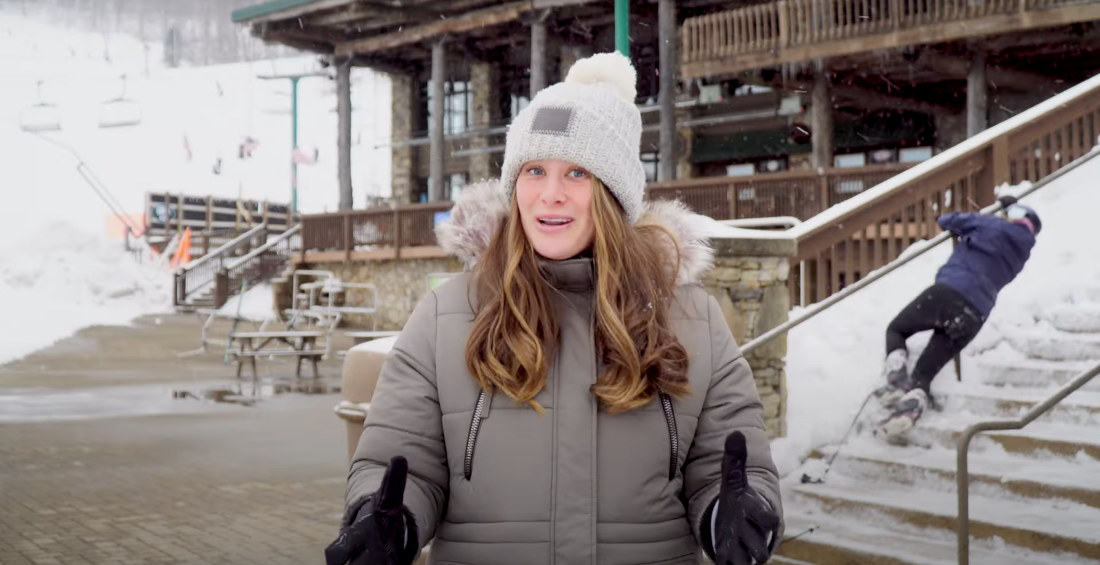 Woman Delivers Ski Resort Snow Report While Skier Struggles Down Stairs In Background